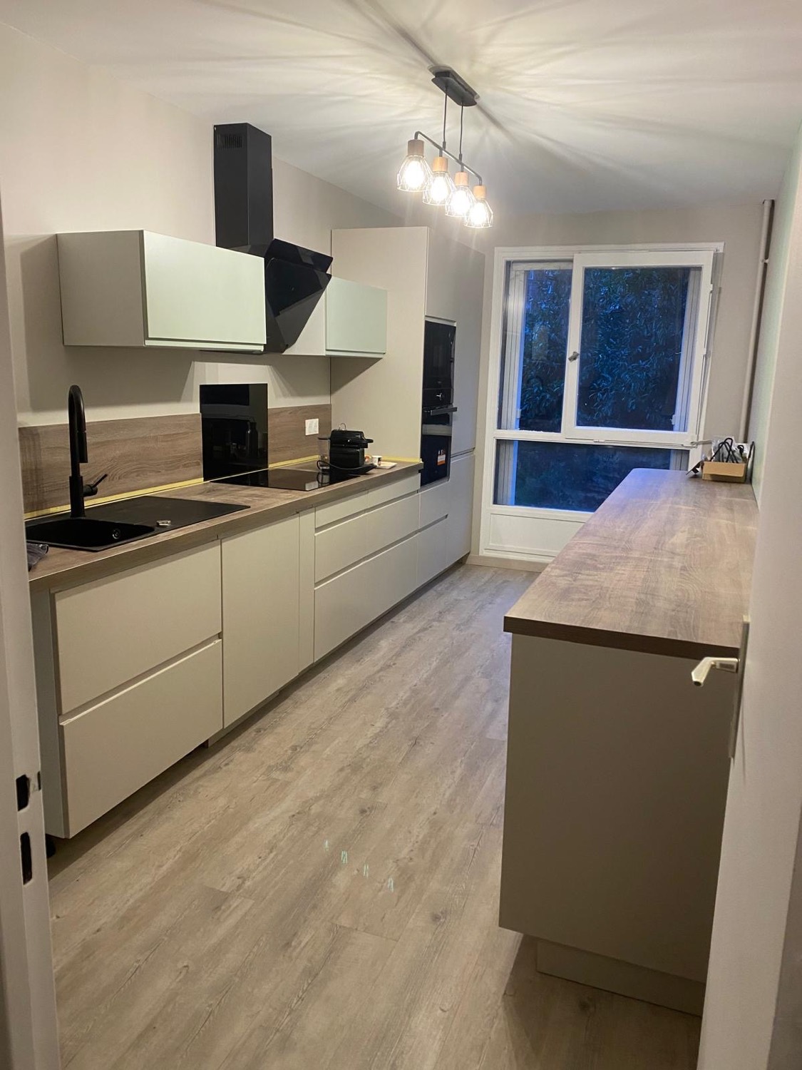 Cuisine équipée moderne aux façades mat taupe, plan de travail et crédence effet bois, hotte et évier noirs, sol stratifié clair — chantier à Deauville