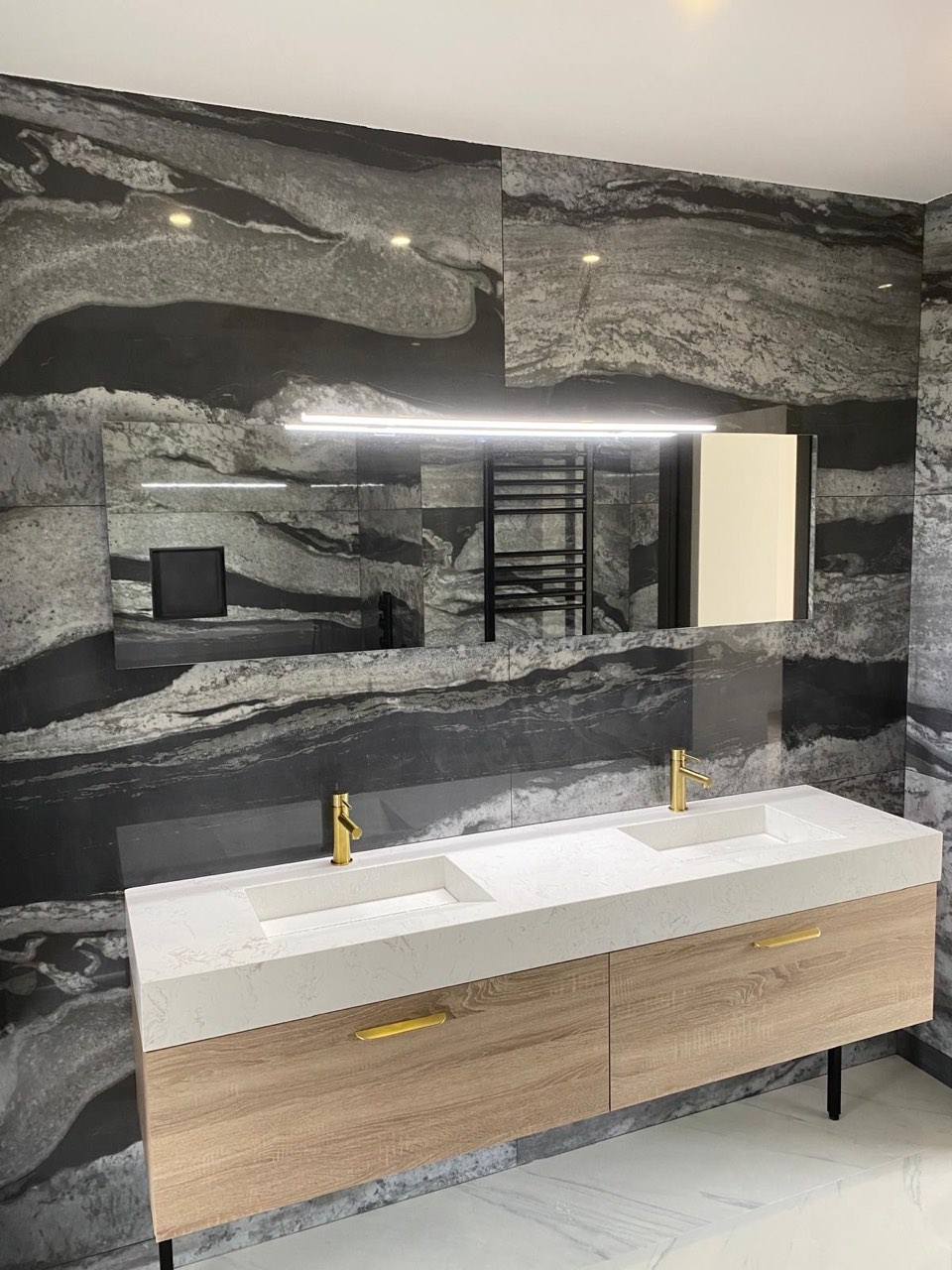 Salle de bain en marbre anthracite veiné avec double vasque en pierre claire sur meuble bois, robinetterie laiton et grand miroir rétroéclairé